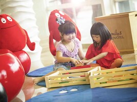 Two children playing with toys in a room with a large red plush toy and wooden play structures.