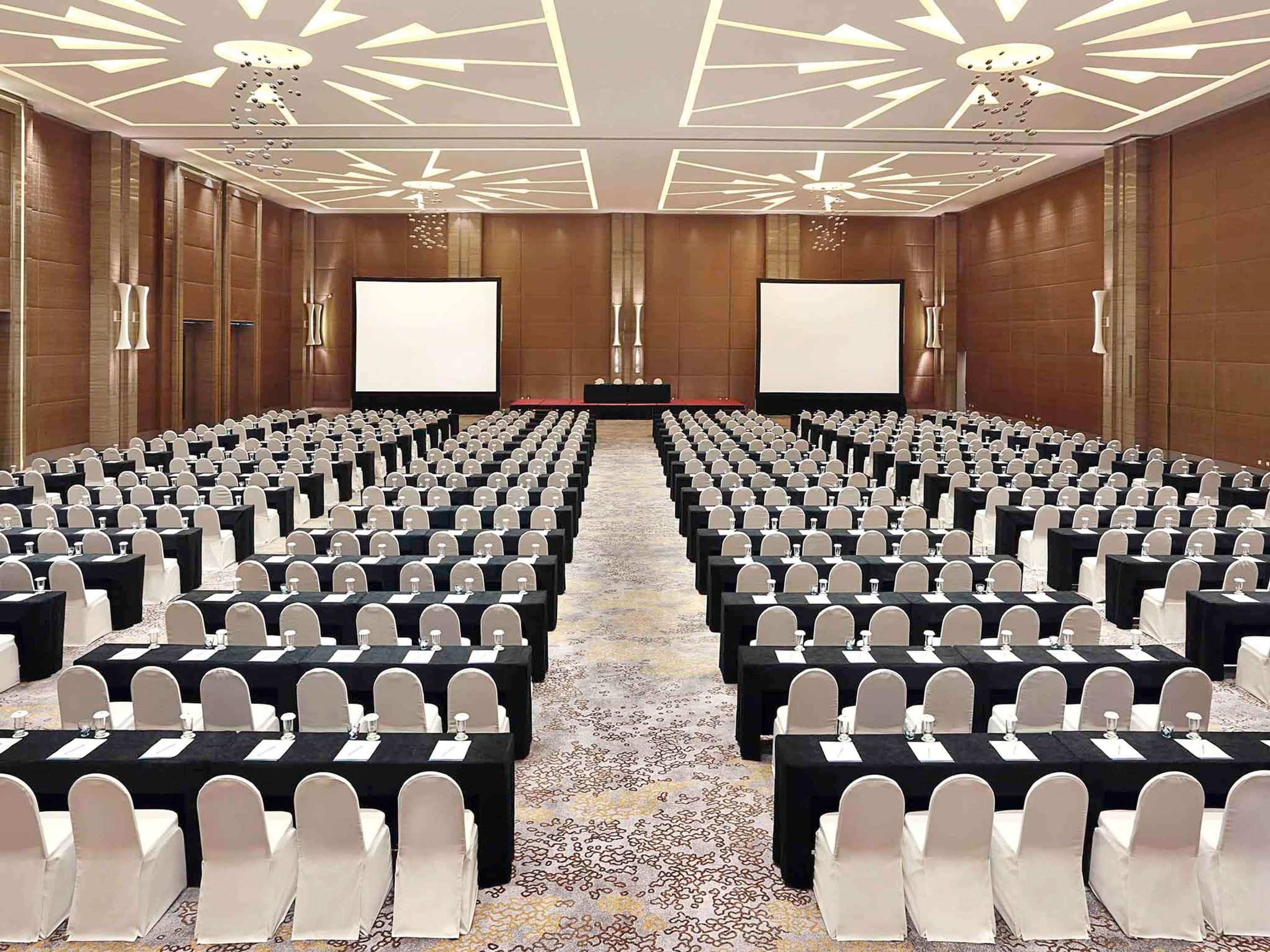 A conference room with rows of tables and chairs, large screens at the front, and a patterned carpet.