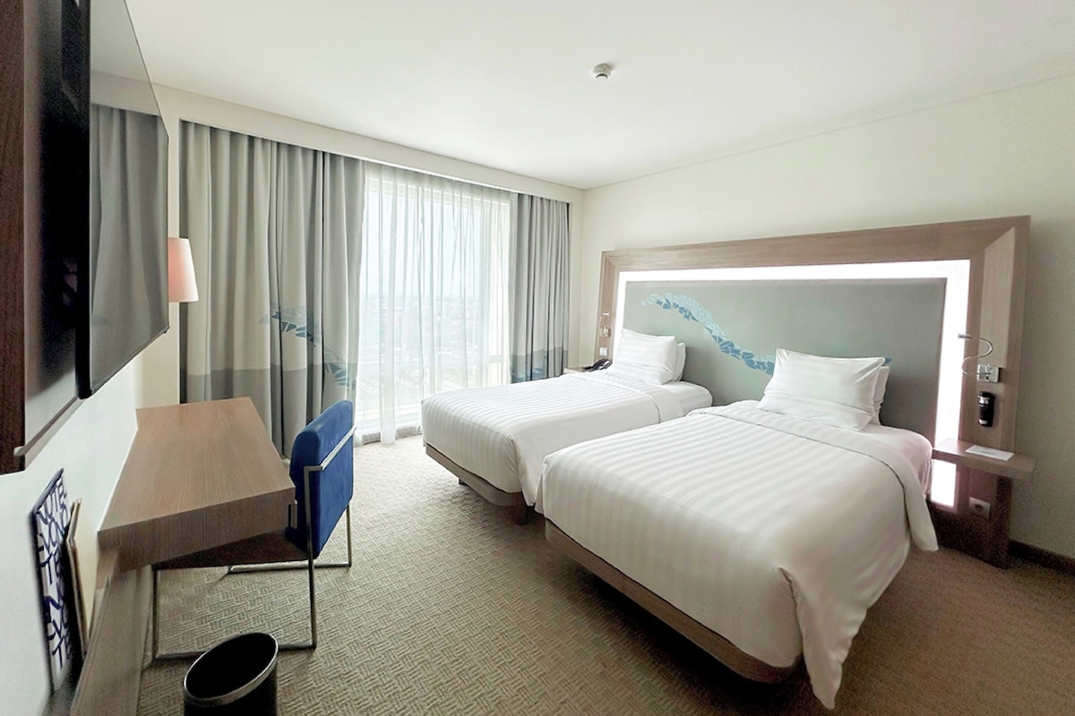 A hotel room with two beds, a desk, a TV, and a large window with curtains.
