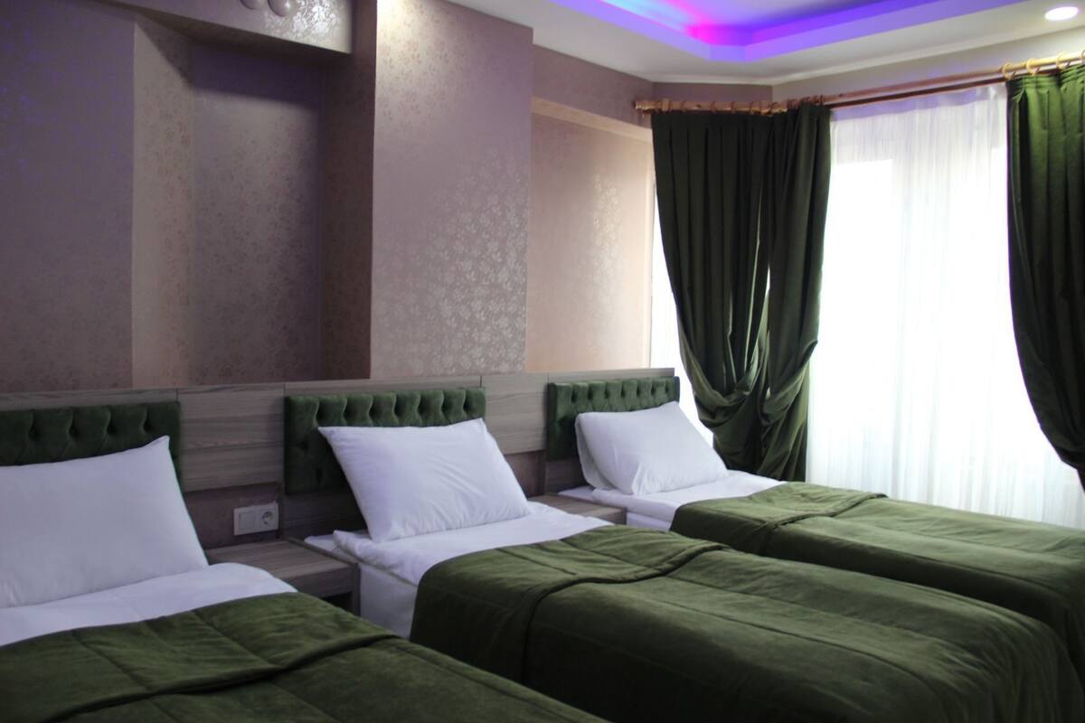 Ein Hotelzimmer mit zwei Betten, einem grünen Kopfteil, weißer Bettwäsche und einem Fenster mit grünen Vorhängen.
