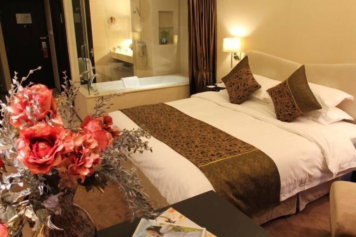 Ein Hotelzimmer mit einem großen Bett, einem Nachttisch mit Blumenvase, einem Spiegel und einem durch eine Glastür einsehbaren Badezimmer.
