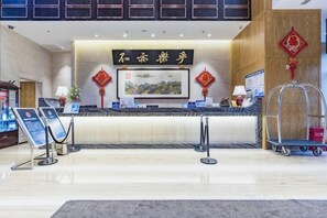A hotel reception area with a counter, informational signs, a framed artwork, and a traditional Chinese decorative element. - Donghuang Kaili Hotel (Beijing)