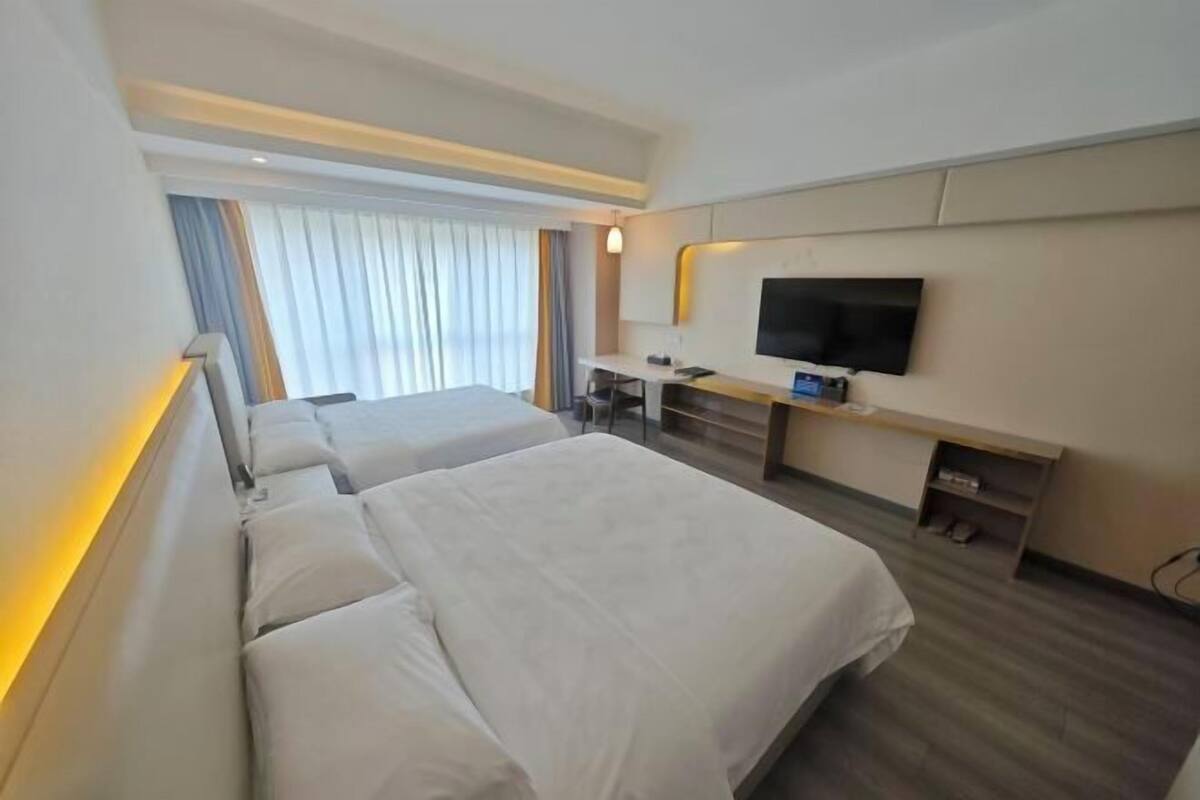 Ein Hotelzimmer mit zwei Betten, einem Schreibtisch, einem Flachbildfernseher und einem großen Fenster mit Vorhängen.