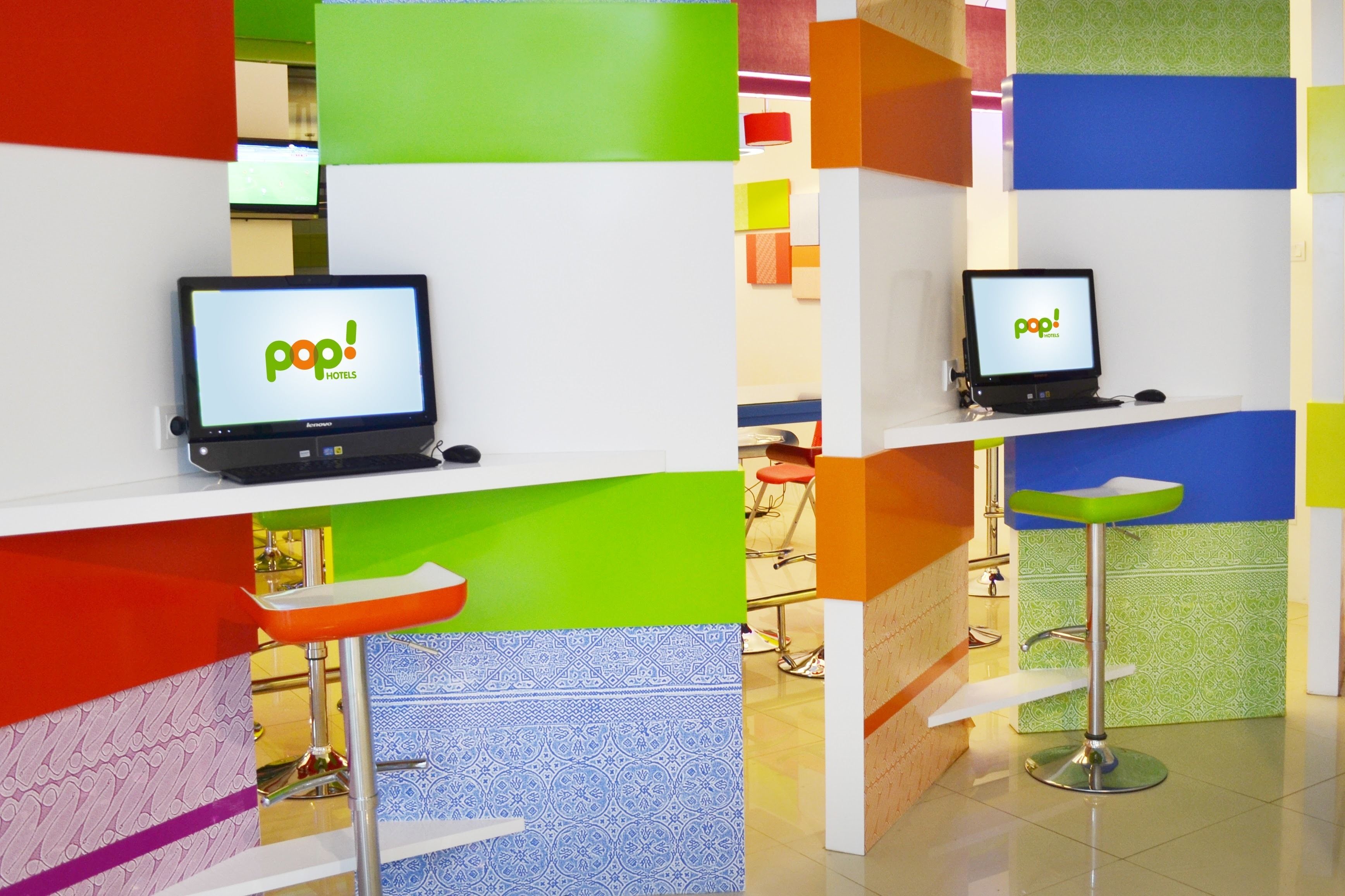Un espace d’accueil d’hôtel avec deux écrans d’ordinateur affichant le logo de « pop! Hotels », un tabouret de bar rouge et un tabouret de bar vert.