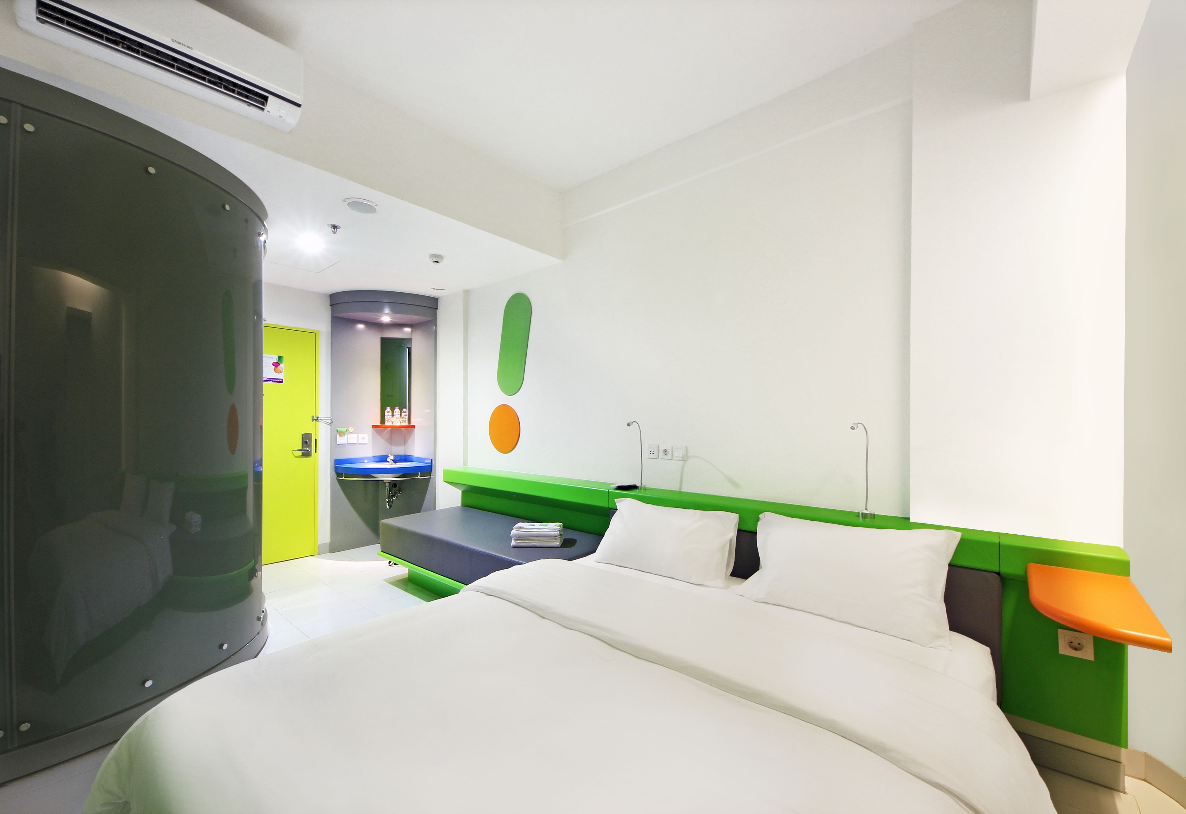 Camera d'albergo con un letto, una parete verde decorativa, una piccola scrivania e un bagno visibile attraverso una porta aperta.