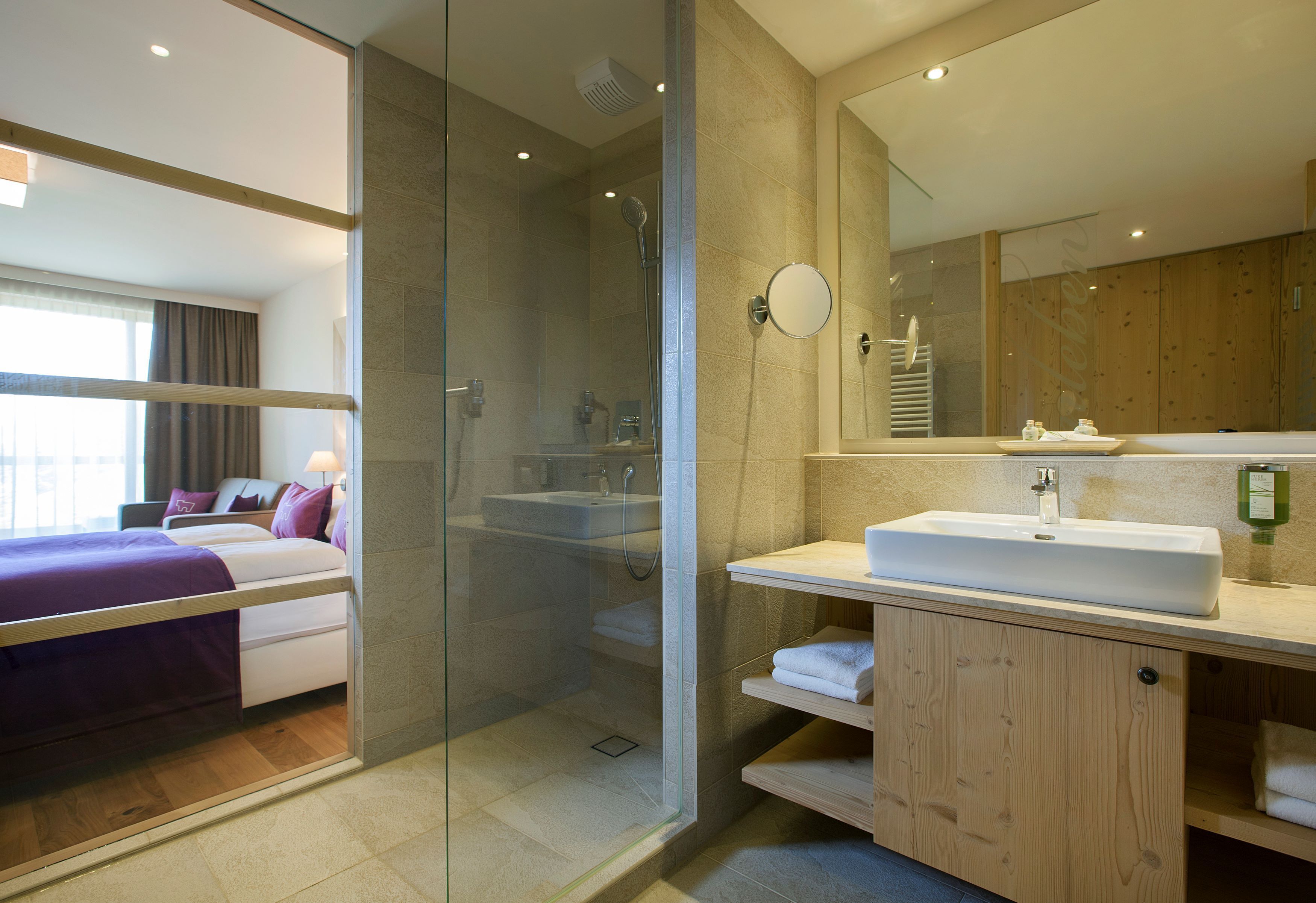 A modern hotel room with a glass-enclosed shower, a large bed with a purple bedspread, a wooden vanity with a white sink, and a large mirror.