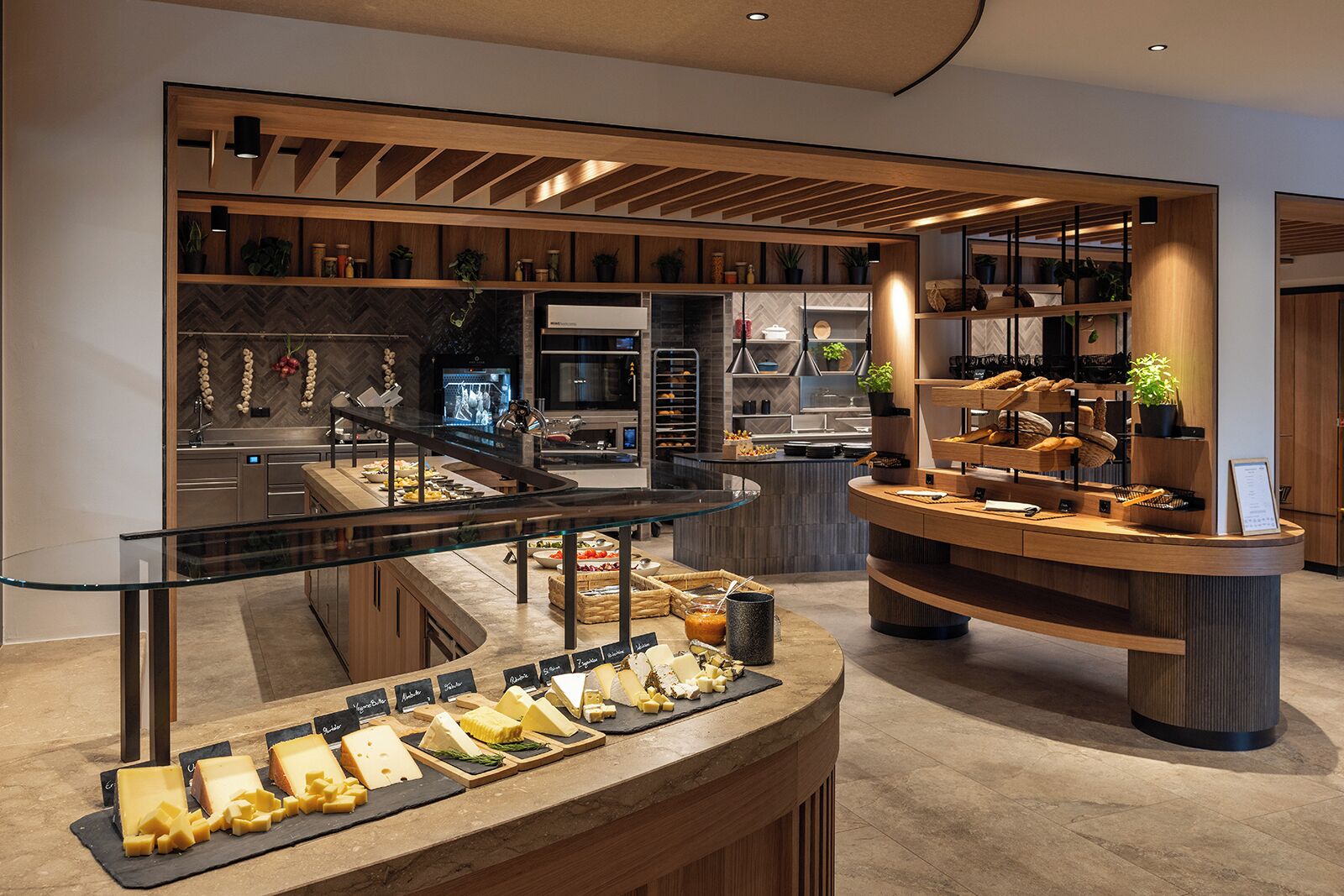 A modern buffet setup with various cheeses and breads, wooden shelves with plants, and a kitchen area in the background.