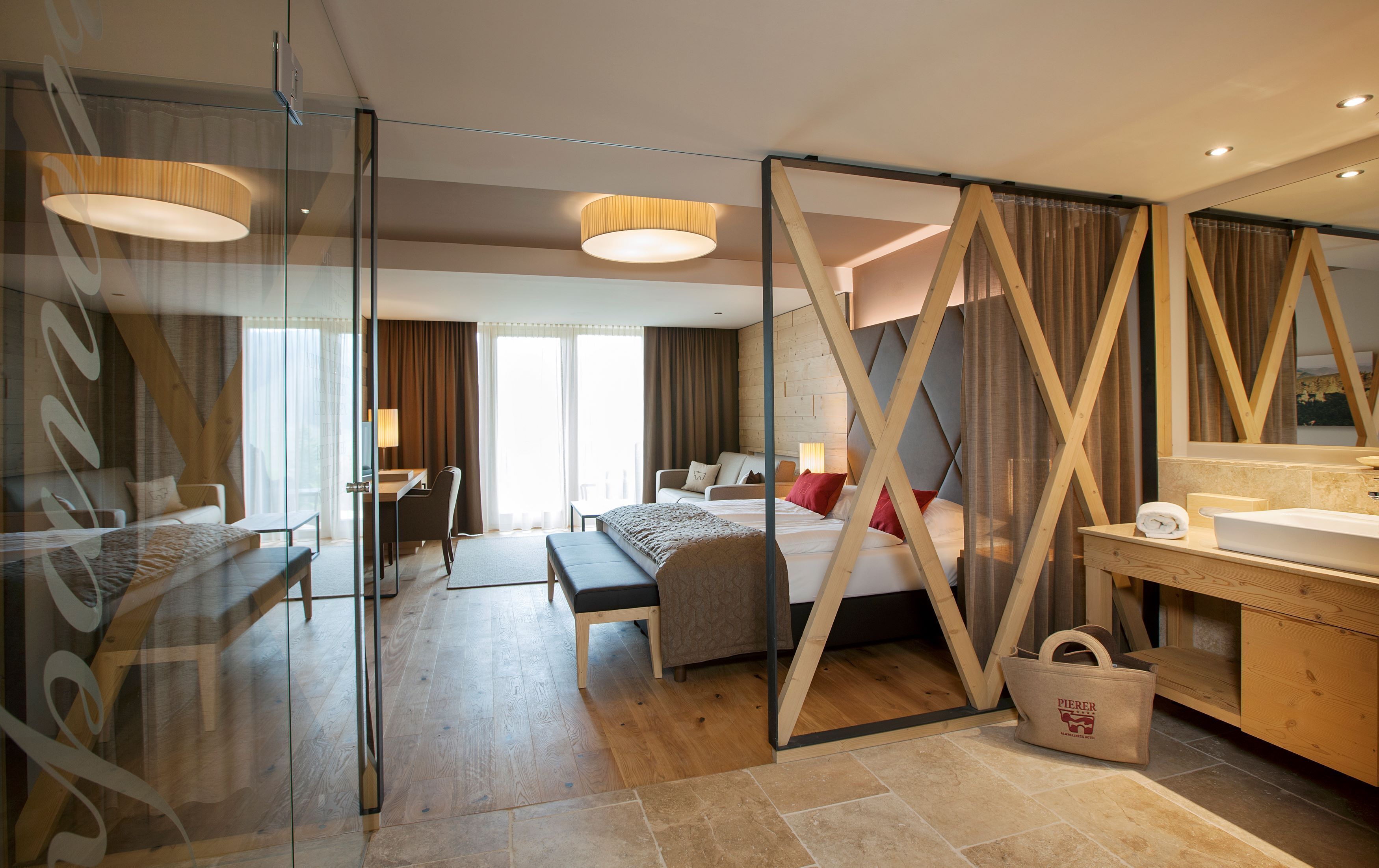 A modern hotel room with a glass partition, a bed, a desk, a sofa, and a bathroom with a wooden vanity.