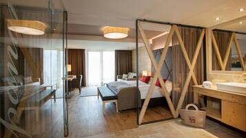 A modern hotel room with a glass partition, a bed, a desk, a sofa, and a bathroom with a wooden vanity.