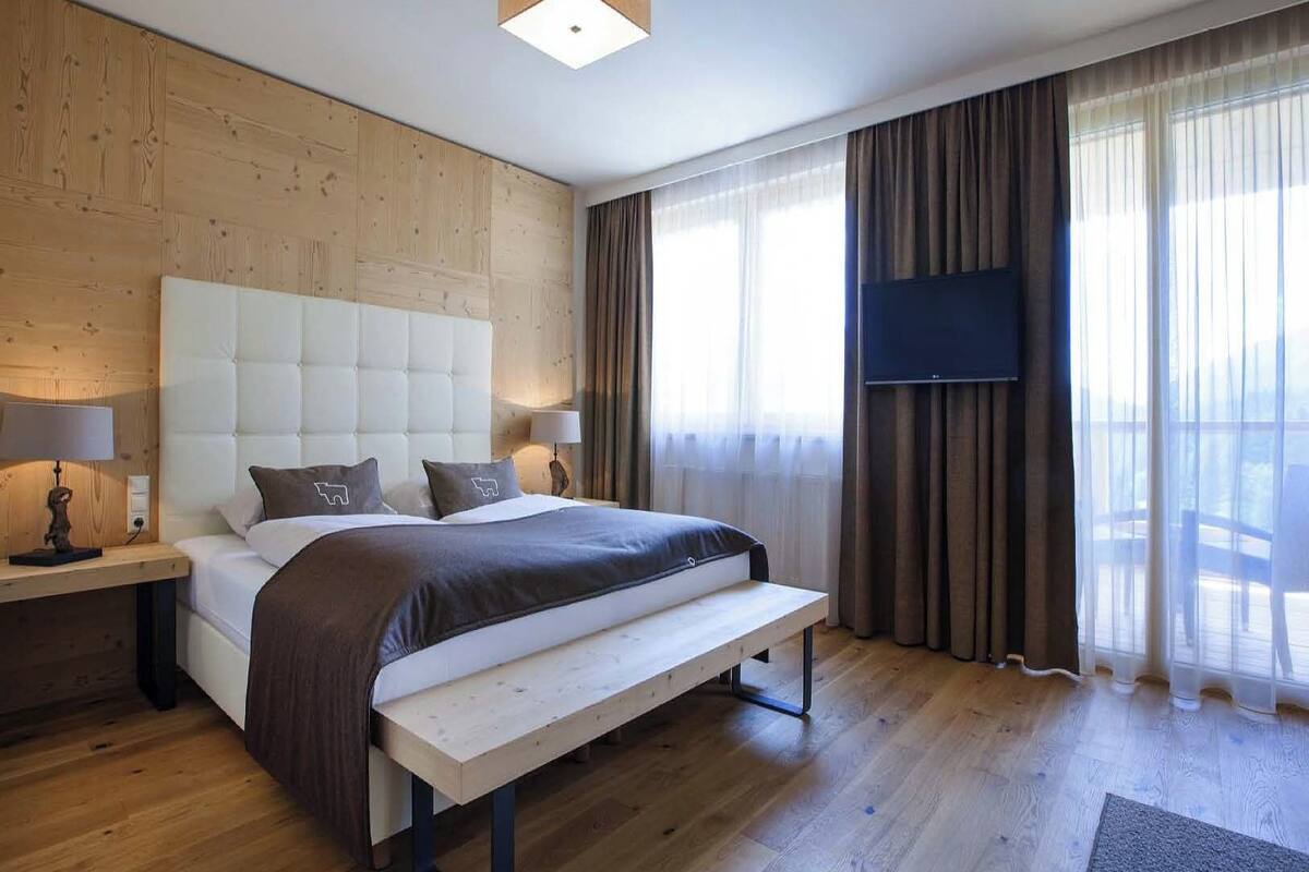 A modern hotel room with a large bed, wooden panel walls, a flat-screen TV, and a bench at the foot of the bed.
