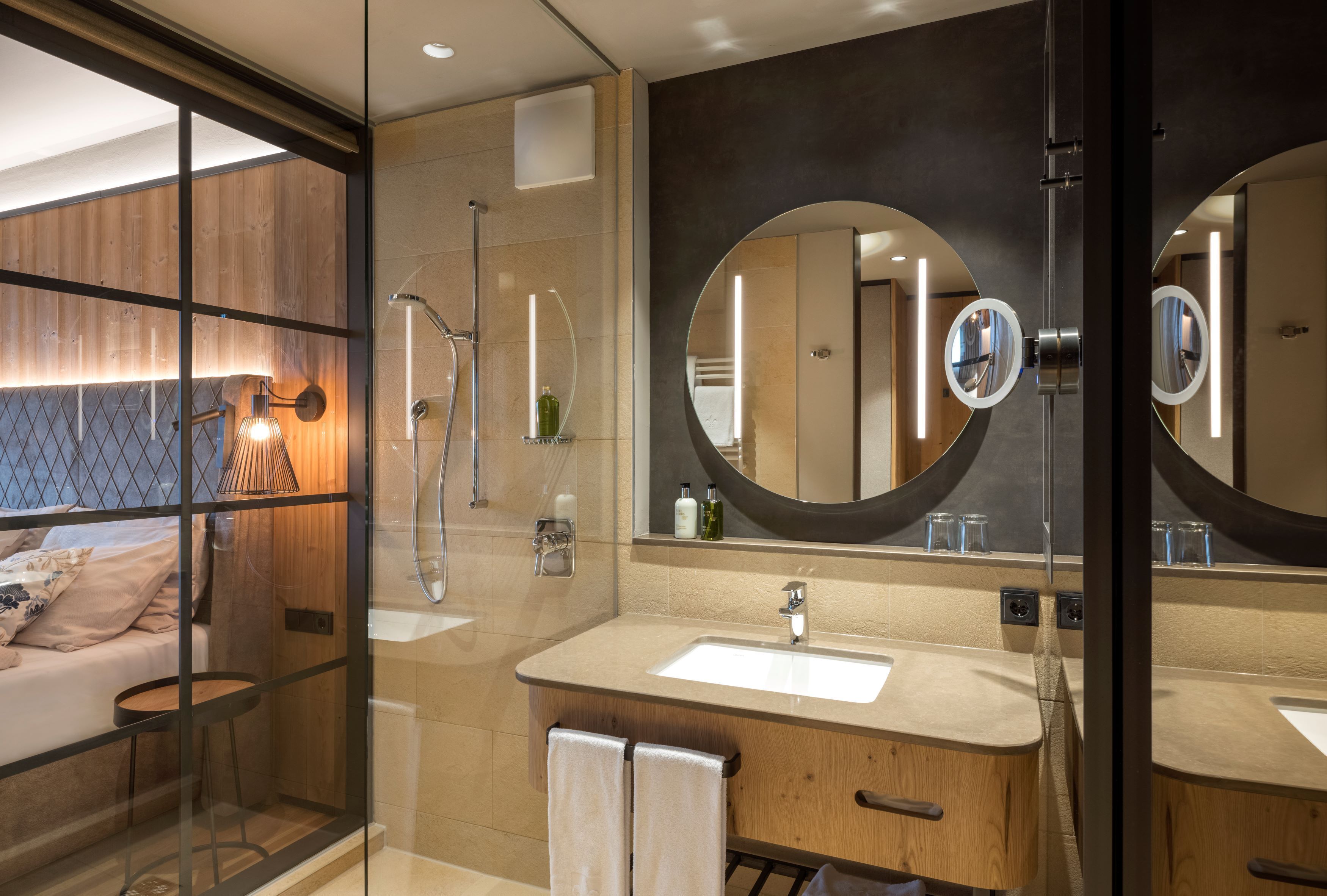 A modern bathroom with a large mirror, a sink, and a shower. There is a glass partition separating the bathroom from a bedroom area.