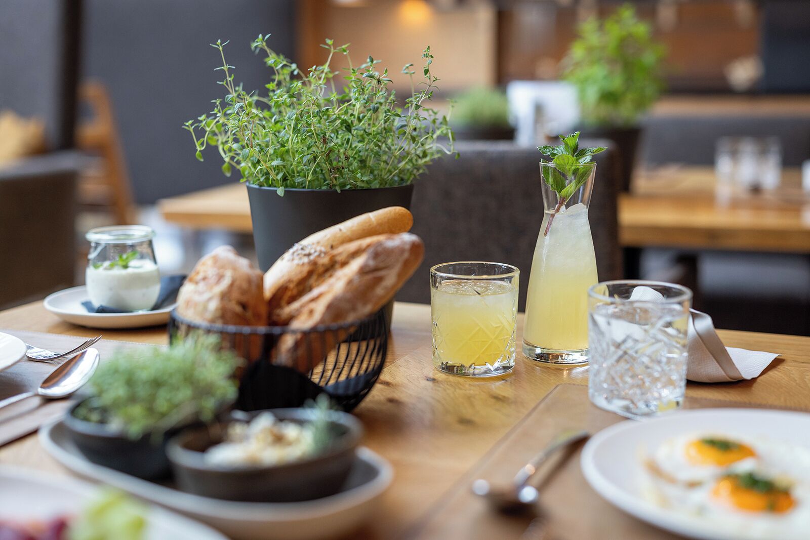 A dining table with a variety of food items, including bread, a bowl of soup, and a plate with eggs. There are also glasses and a pitcher of a yellow beverage.