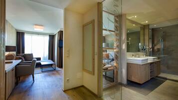 A modern hotel room with a wooden floor, a sofa, a TV, a glass partition, and a bathroom with a shower and sink.