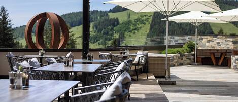An outdoor patio with tables and chairs, a stone wall, and a view of a forested hillside.