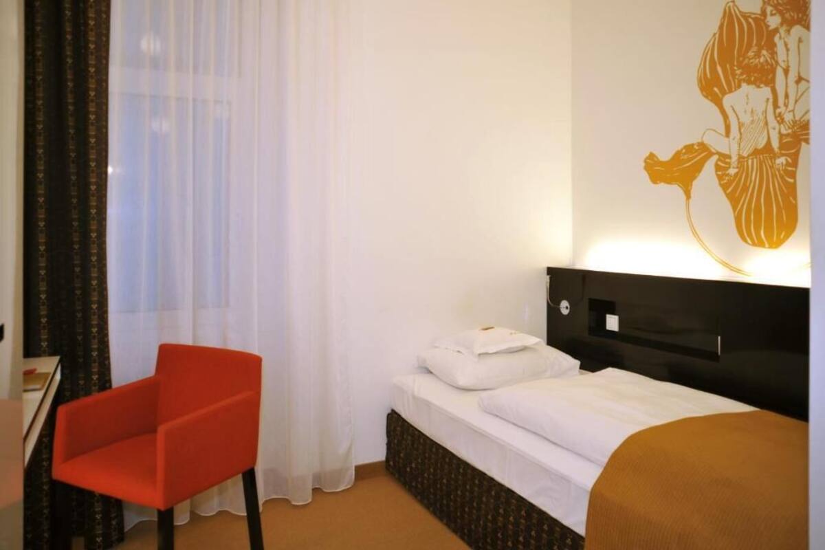 A hotel room with a bed, a red chair, a desk, and a wall with a decorative painting.