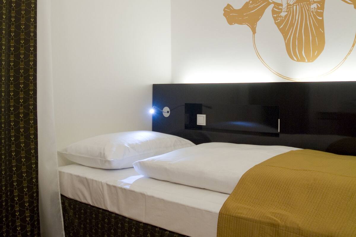 A hotel room with a bed, a headboard, a wall-mounted light, and a curtain.