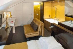 Quarto de hotel compacto com sofá-cama, uma pequena mesa de cabeceira com luminária, uma cama de solteiro e banheiro com pia e chuveiro.