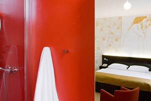 Quarto de hotel moderno com parede de destaque vermelha, cortina de chuveiro branca e cama com roupa de cama branca e manta dourada.