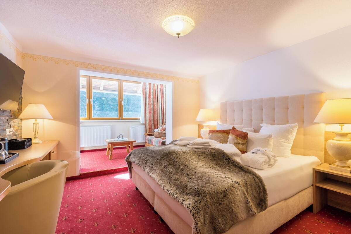 A hotel room with a bed, two bedside tables, a chair, a small table, and a window with curtains.