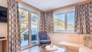 A modern living room with a sofa, a glass coffee table, and a chair. Large windows offer a view of a mountainous landscape. - Hotel Alpenkönigin Alpine Luxury   (See)