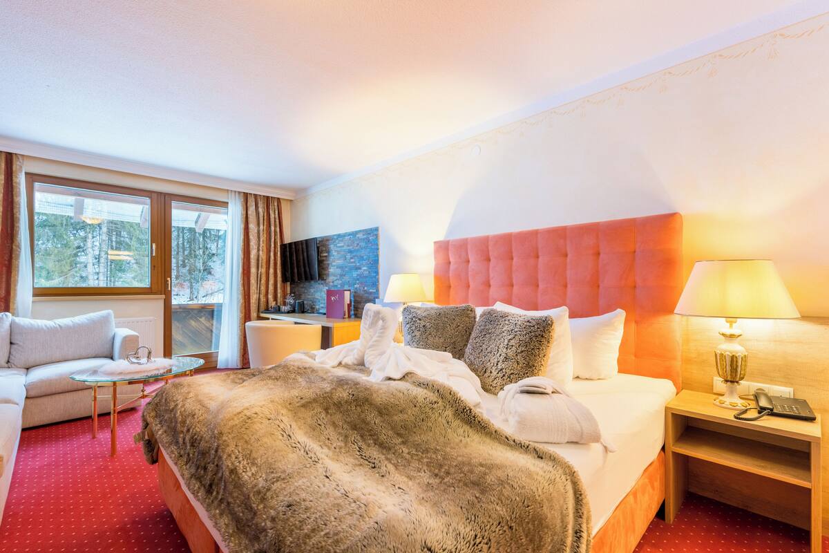 A hotel room with a large bed, a sofa, a small table, and a view of the city through the window.