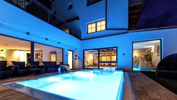 A modern indoor swimming pool with a glass enclosure, surrounded by lounge seating and a covered patio area. - Hotel Alpenkönigin Alpine Luxury   (See)