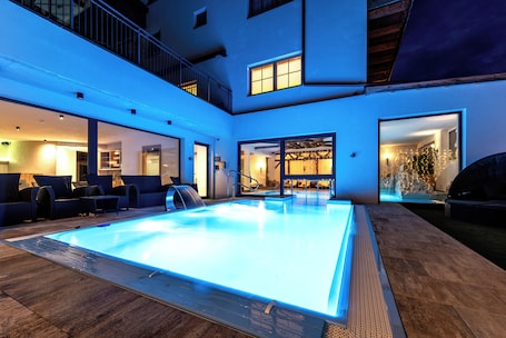 A modern indoor swimming pool with a glass enclosure, surrounded by lounge seating and a covered patio area.