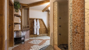 A rustic bathroom with a stone mosaic floor, wooden shelves with towels, and a shower area with stone walls. - Hotel Alpenkönigin Alpine Luxury   (See)