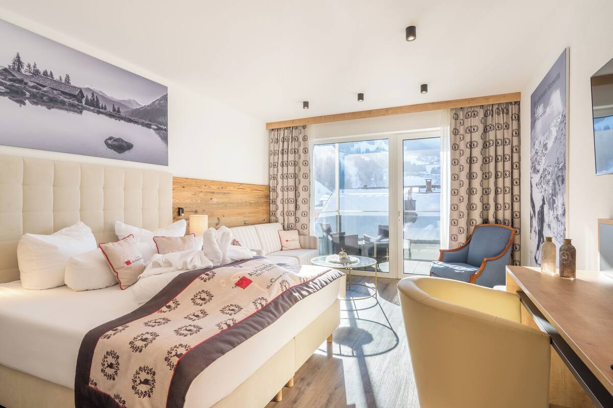 A hotel room with a large bed, a sofa, a dining table, and a balcony with a view of the mountains.