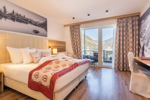 A hotel room with a large bed, a desk, and a view of mountains through the window. - Hotel Alpenkönigin Alpine Luxury   (See)