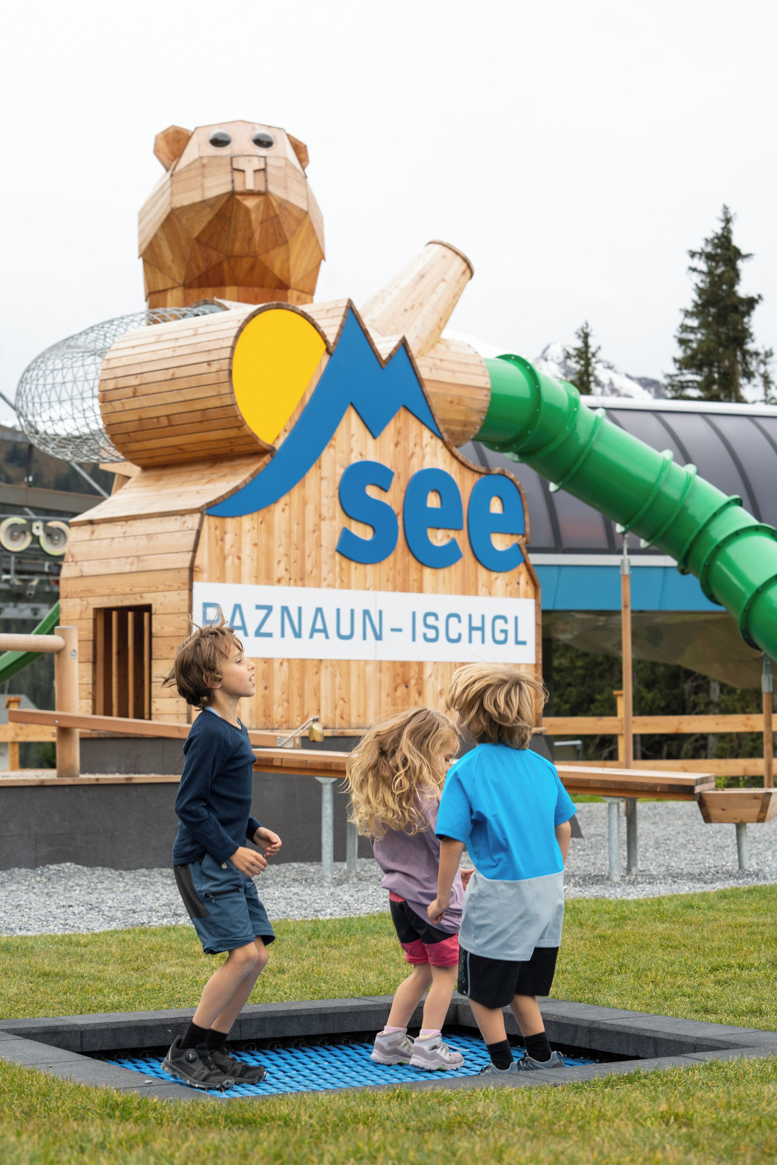 Estructura de madera con diseño de oso y un tobogán, situada en un área al aire libre con césped y parque infantil.
