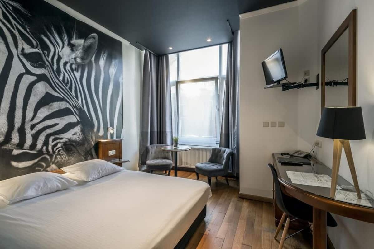 Habitación de hotel con un gran mural de cebra, una cama, un escritorio con computadora, una silla, un televisor y una ventana con cortinas.