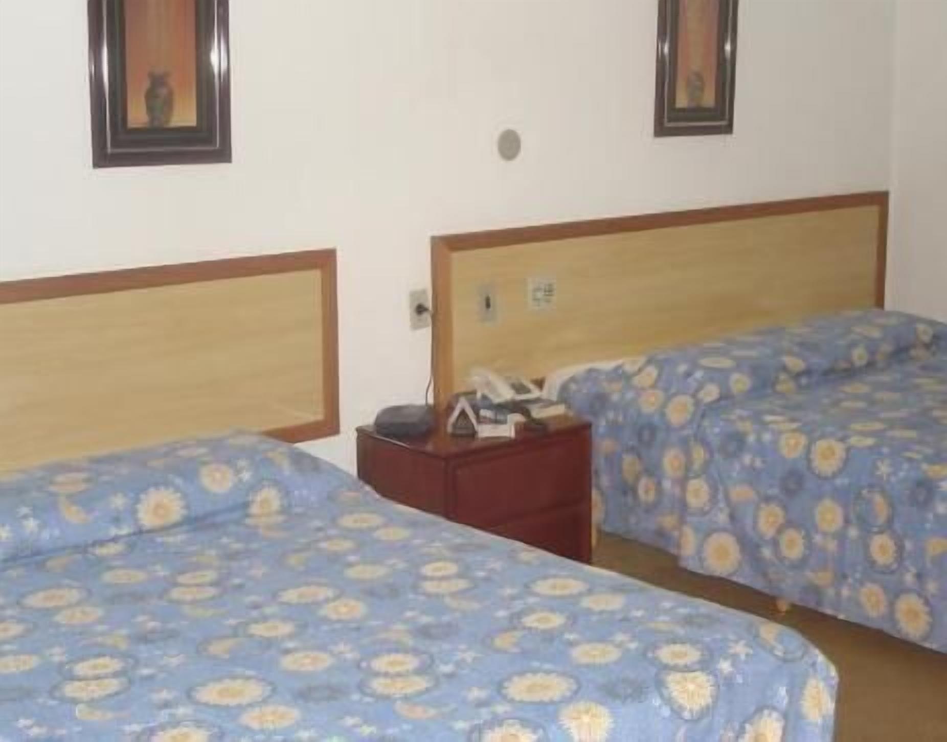 Habitación de hotel con dos camas, cada una con cabecera de madera y ropa de cama con estampado floral azul. Entre las camas hay una mesita de noche con una lámpara y un teléfono.
