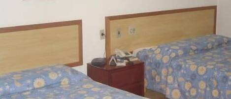 Habitación de hotel con dos camas, cada una con cabecera de madera y ropa de cama con estampado floral azul. Entre las camas hay una mesita de noche con una lámpara y un teléfono.