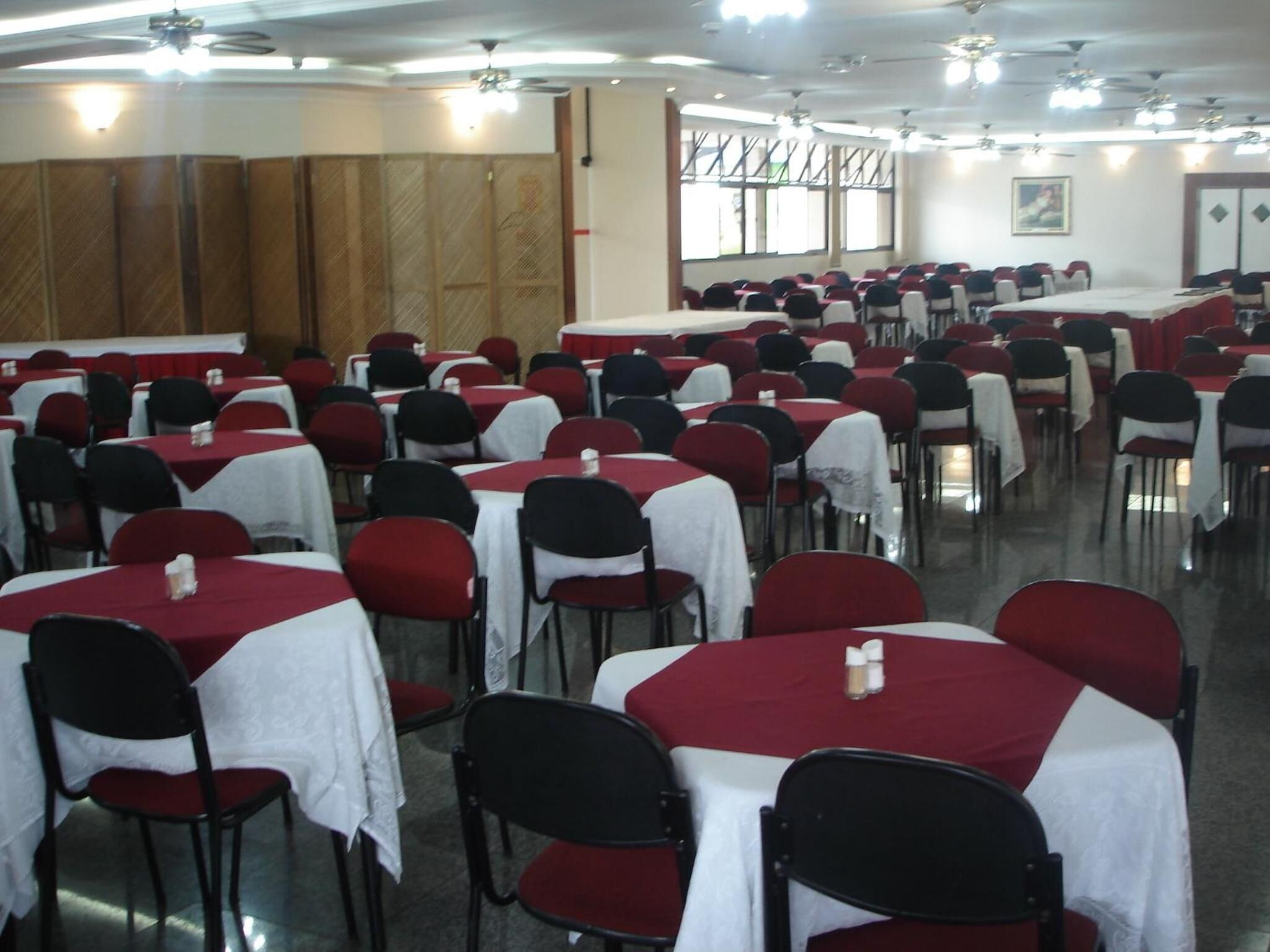 Un salón amplio con filas de mesas y sillas, algunas cubiertas con manteles blancos y otras con fundas rojas.