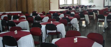 Un salón amplio con filas de mesas y sillas, algunas cubiertas con manteles blancos y otras con fundas rojas.