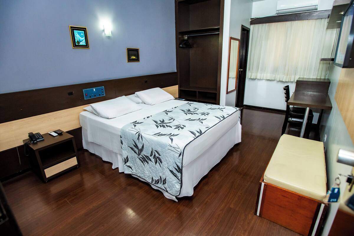 A hotel room with a bed, bedside table, wardrobe, and a small desk with a chair.