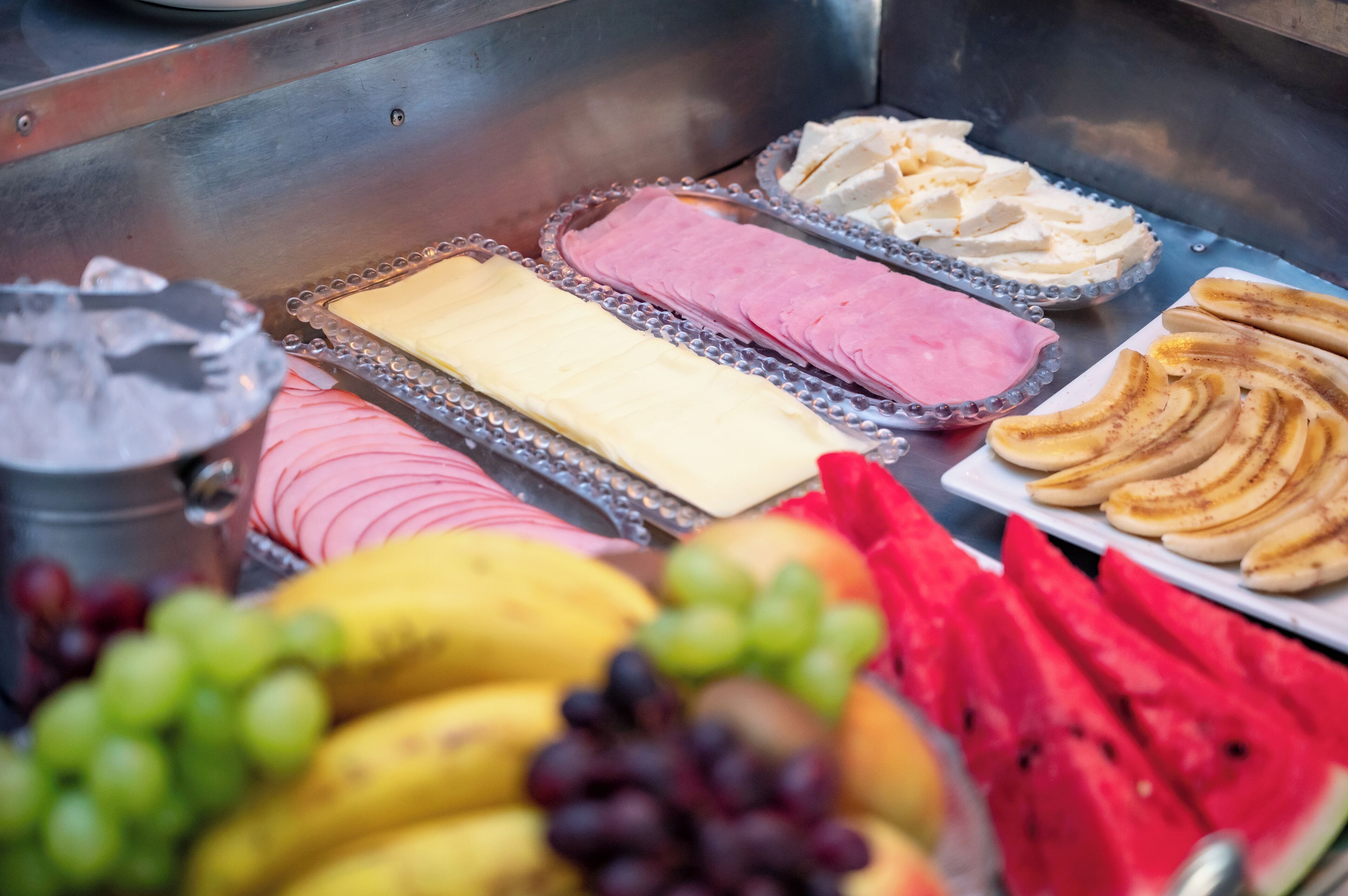 Un bufé con diversas opciones de comida, incluyendo plátanos en rodajas, sandía, uvas, y diferentes tipos de quesos y embutidos.