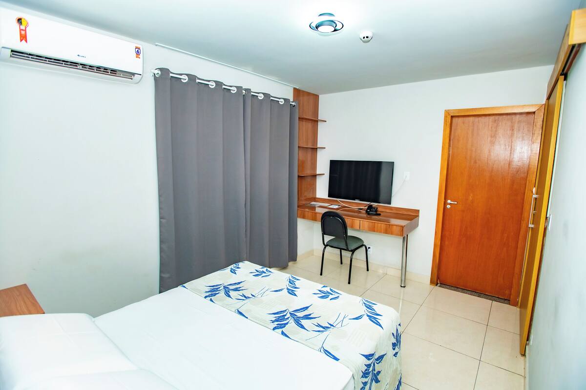 A hotel room with a bed, a desk with a computer, a chair, an air conditioning unit, a wooden door, and a television.