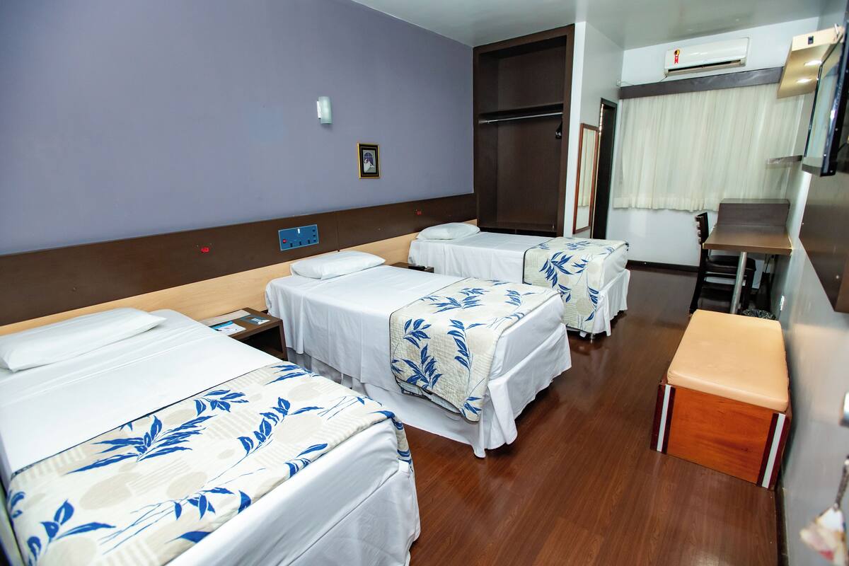 A hotel room with three beds, a wooden bench, a desk, and an air conditioning unit.