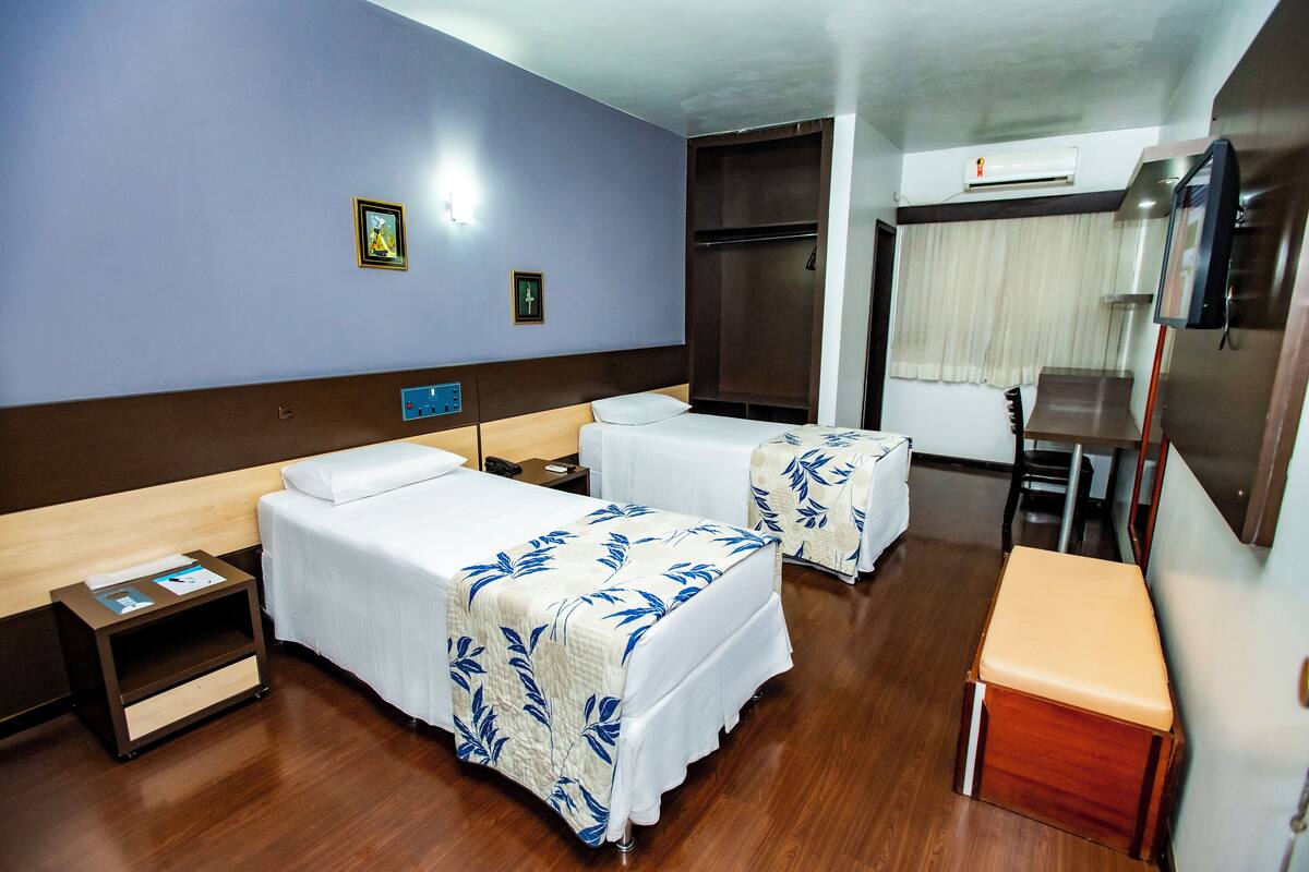 A hotel room with two beds, a small table, a bench, a wardrobe, and a television.