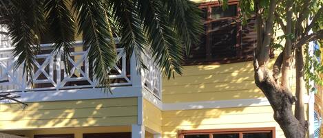 A two-story yellow house with a white balcony, a small porch, and a tree in the front yard.