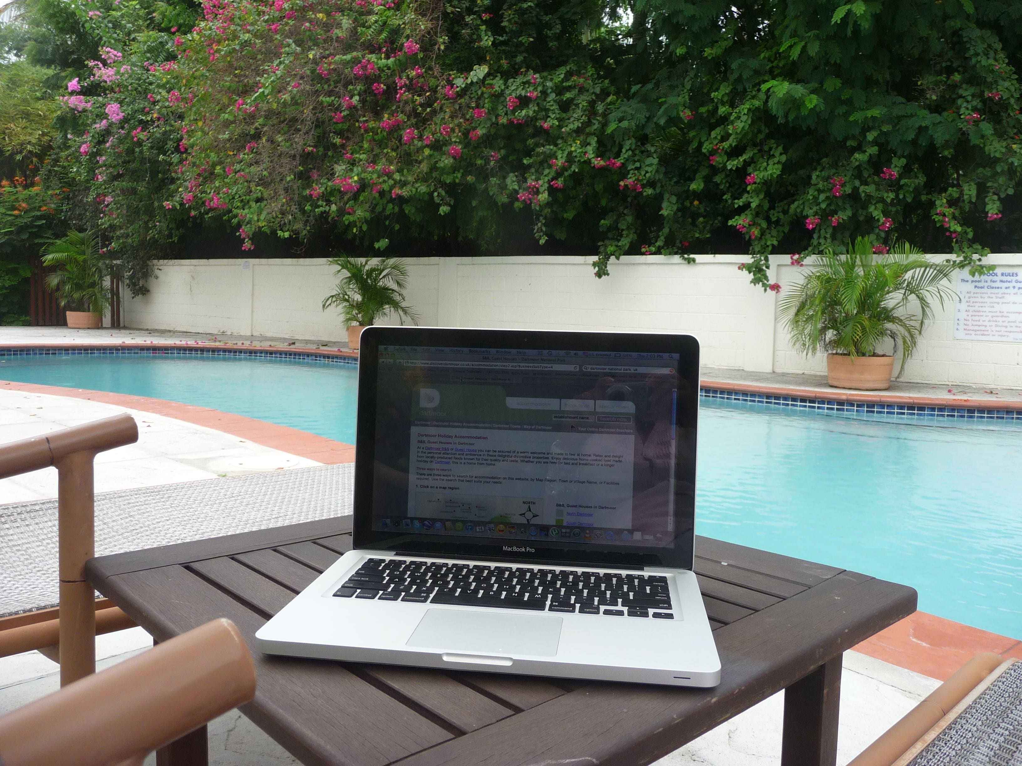 Una computadora portátil sobre una mesa, cerca de una piscina con vista a un jardín y un muro con texto.
