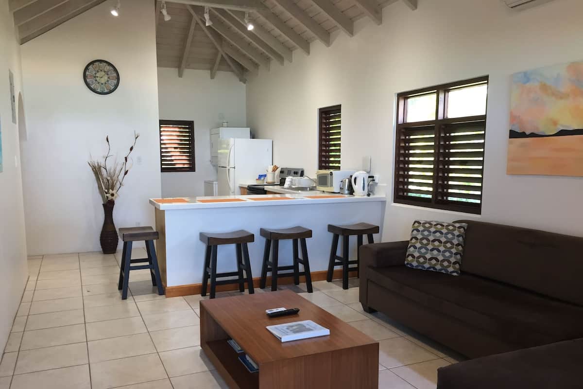 A modern living room with a brown sofa, a wooden coffee table, and a kitchen area with a white refrigerator and bar stools.