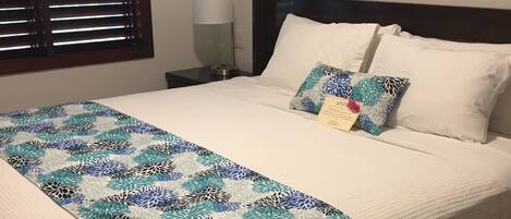 A neatly made bed with a floral-patterned bedspread, a bedside lamp, and a window with blinds.