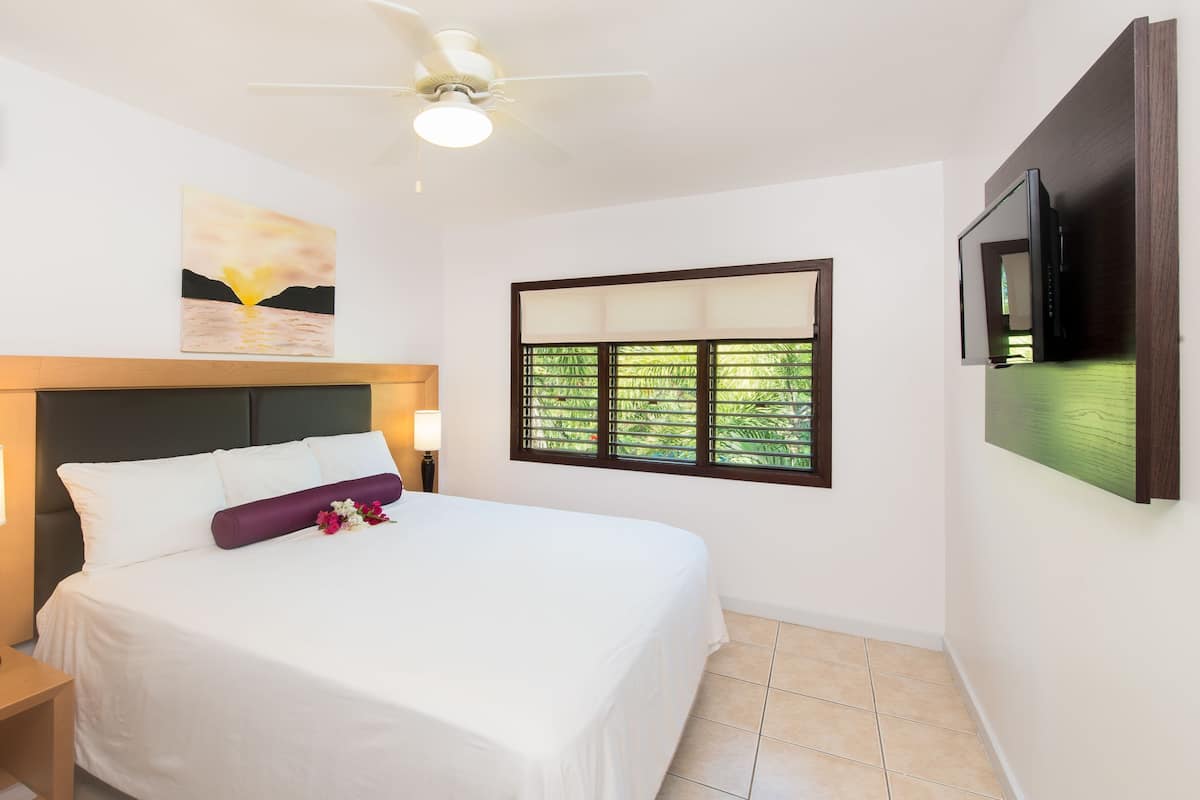 A hotel room with a bed, bedside lamps, a ceiling fan, a window with blinds, and a TV mounted on the wall.