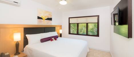 A hotel room with a bed, bedside lamps, a ceiling fan, a window with blinds, and a TV mounted on the wall.