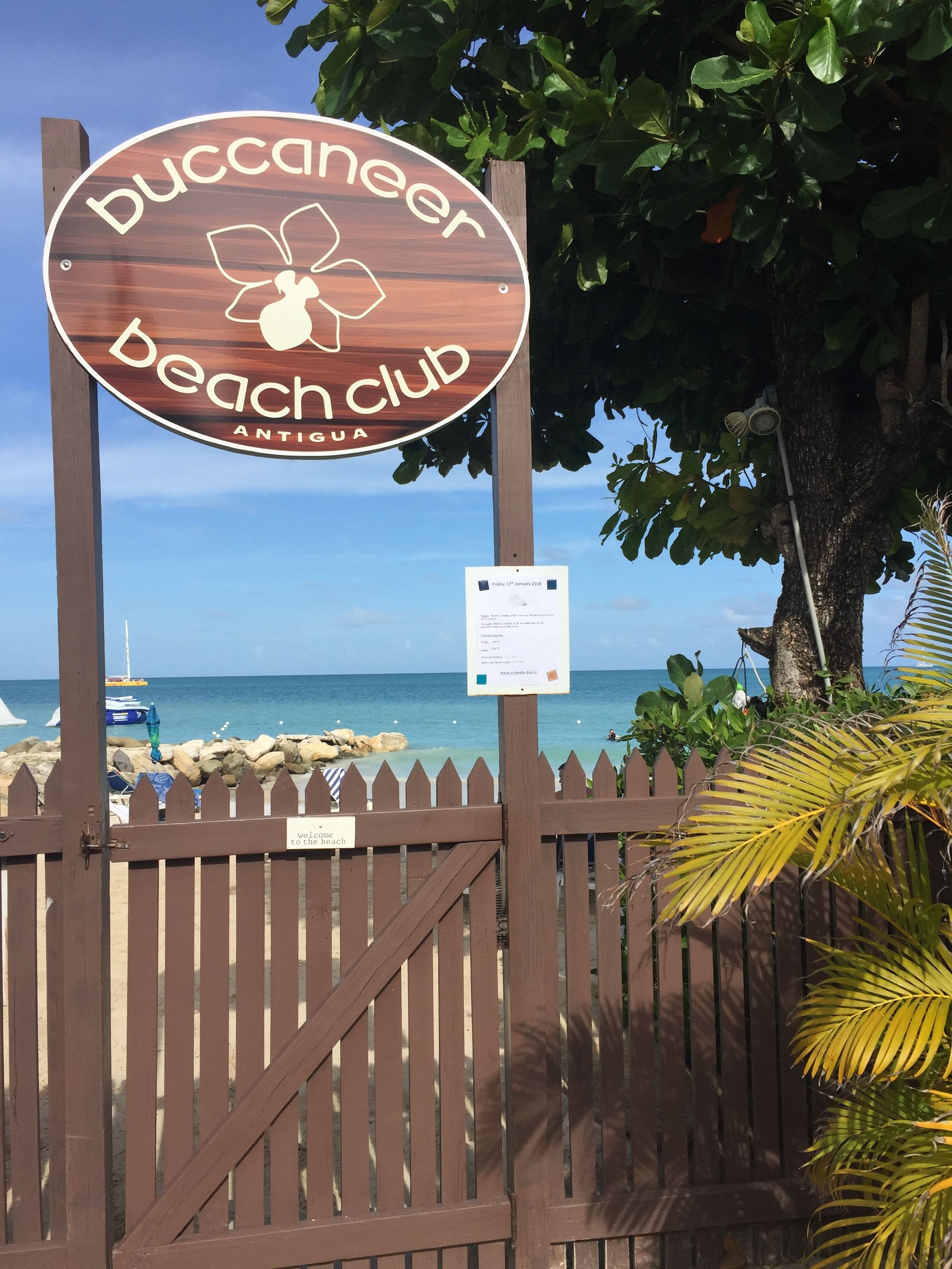 A wooden sign for Buccaneer Beach Club with a palm tree and a wooden fence in the foreground.