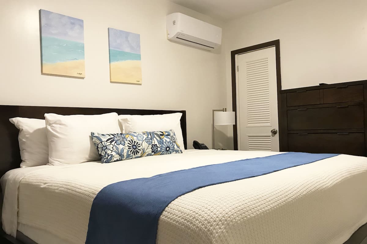 A neatly made bed with white and blue bedding, a floral pillow, and a blue throw blanket. There are two wall-mounted paintings depicting beach scenes.