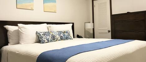 A neatly made bed with white and blue bedding, a floral pillow, and a blue throw blanket. There are two wall-mounted paintings depicting beach scenes.